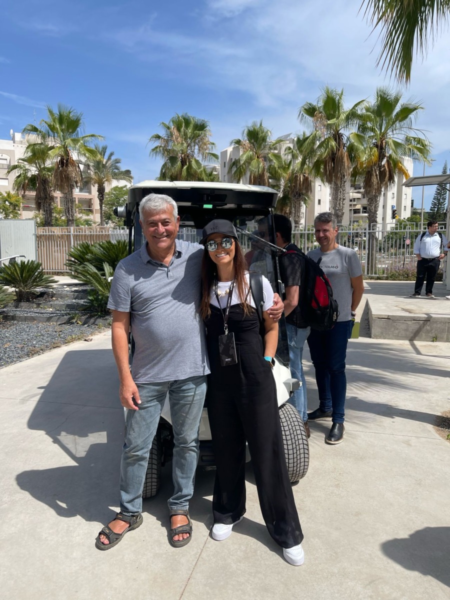 Homem idoso, de cabelos grisalhos, de calça jeans e camisa polo cinza com o braço sobre os ombros de uma mulher de boné, óculos escuros, cabelos soltos e lisos, vestindo um macacão preto e uma camiseta branca, com o celular em um cordão no pescoço e uma mão no bolso, em frente a um carro de golfe autônomo, em Israel. 